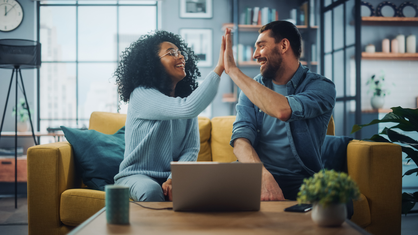 Diverse Multiethnic Couple are Sitting on a Couch Sofa in Stylish Living Room and Choosing Items to Buy Online with Laptop Computer, Give High Five. Friends or Colleagues are Discuss Work Projects